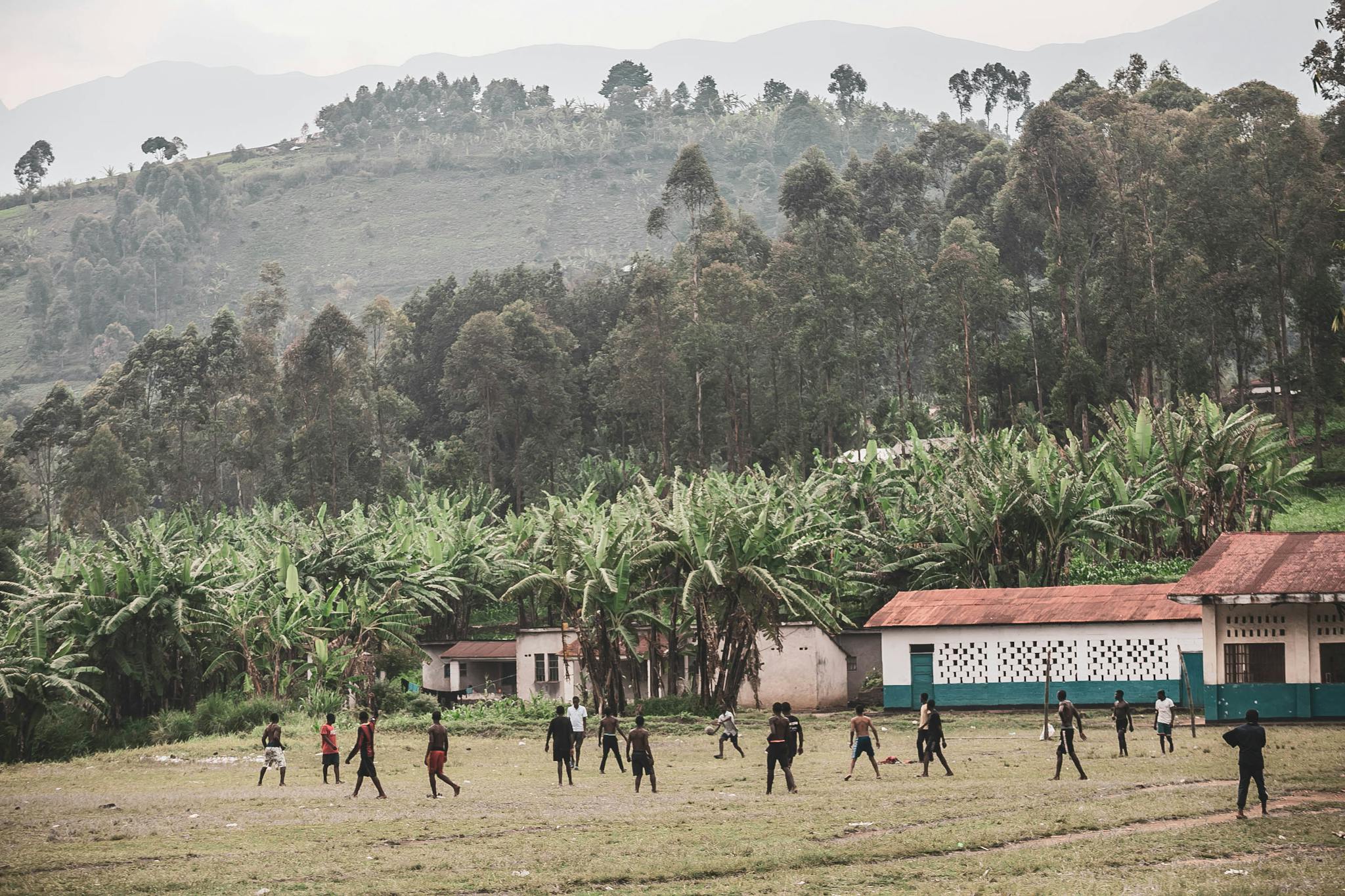 Children play soccer on a grassy field surrounded by lush African landscape and small village buildings.