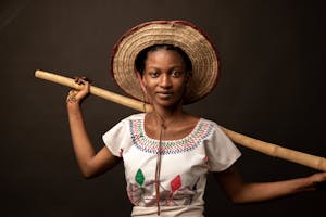 Studio portrait of a woman in a straw hat holding a bamboo stick, wearing an embroidered blouse.