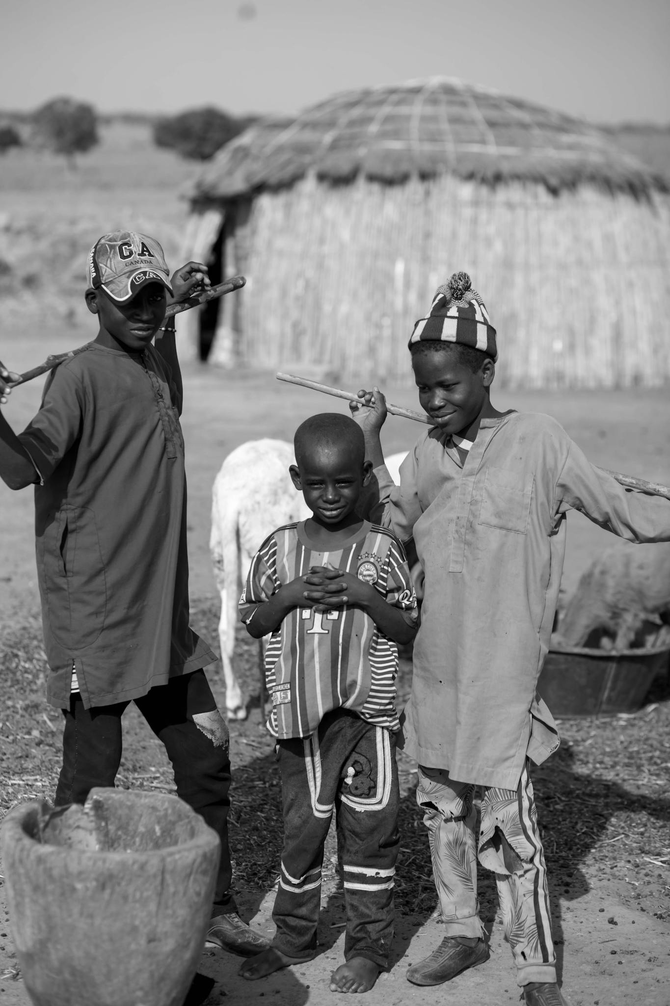 Three children in traditional attire standing outside a hut in a rural area.